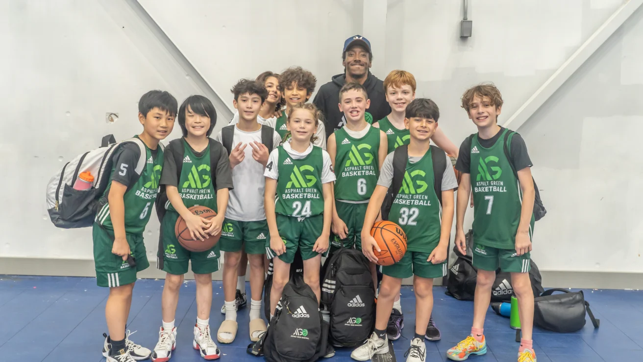 A youth basketball team in green uniforms, proud of their basketball skills, poses for a group photo with their coach inside a gymnasium, holding bags and a basketball.