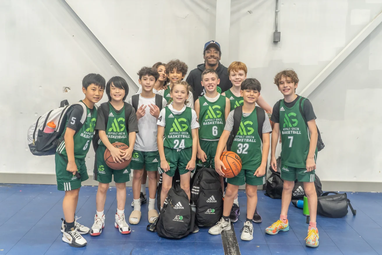 A youth basketball team in green uniforms, proud of their basketball skills, poses for a group photo with their coach inside a gymnasium, holding bags and a basketball.