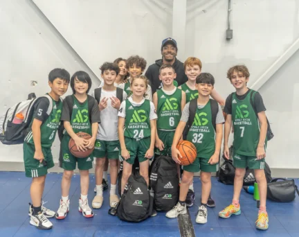 A youth basketball team in green uniforms, proud of their basketball skills, poses for a group photo with their coach inside a gymnasium, holding bags and a basketball.