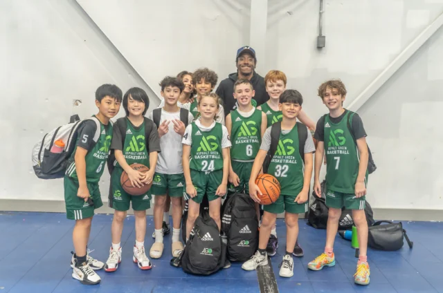 A youth basketball team in green uniforms, proud of their basketball skills, poses for a group photo with their coach inside a gymnasium, holding bags and a basketball.