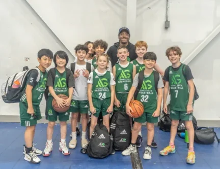 A youth basketball team in green uniforms, proud of their basketball skills, poses for a group photo with their coach inside a gymnasium, holding bags and a basketball.
