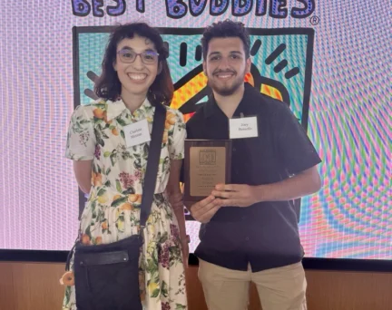 Two people stand in front of a "Best Buddies" sign. One holds a plaque, and both wear nametags and smile at the camera.