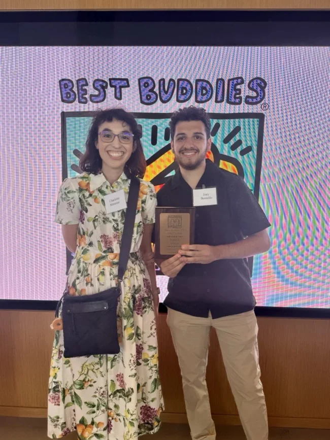 Two people stand in front of a "Best Buddies" sign. One holds a plaque, and both wear nametags and smile at the camera.