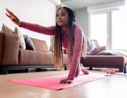 Woman in athletic clothes balances on hands and knees, extending one arm and opposite leg, practicing at-home exercises on a pink yoga mat in a living room.