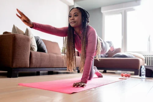 Woman in athletic clothes balances on hands and knees, extending one arm and opposite leg, practicing at-home exercises on a pink yoga mat in a living room.