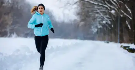 A person wearing a light blue jacket, black leggings, gloves, and a headband runs on a snow-covered path through a winter landscape.