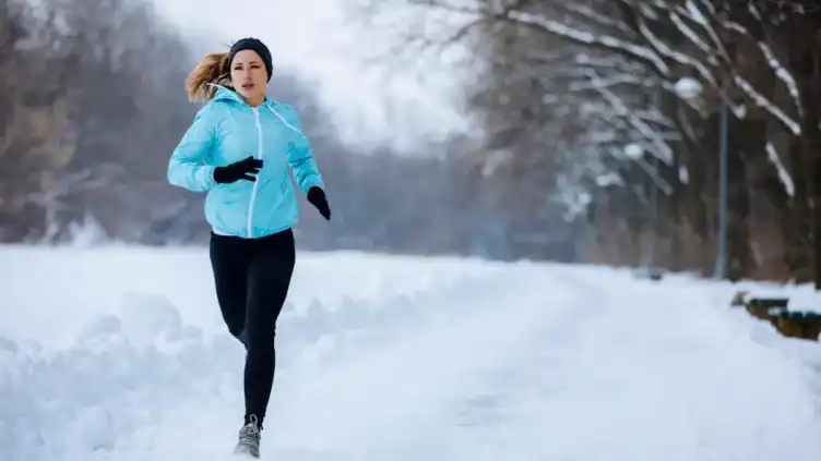 A person wearing a light blue jacket, black leggings, gloves, and a headband runs on a snow-covered path through a winter landscape.