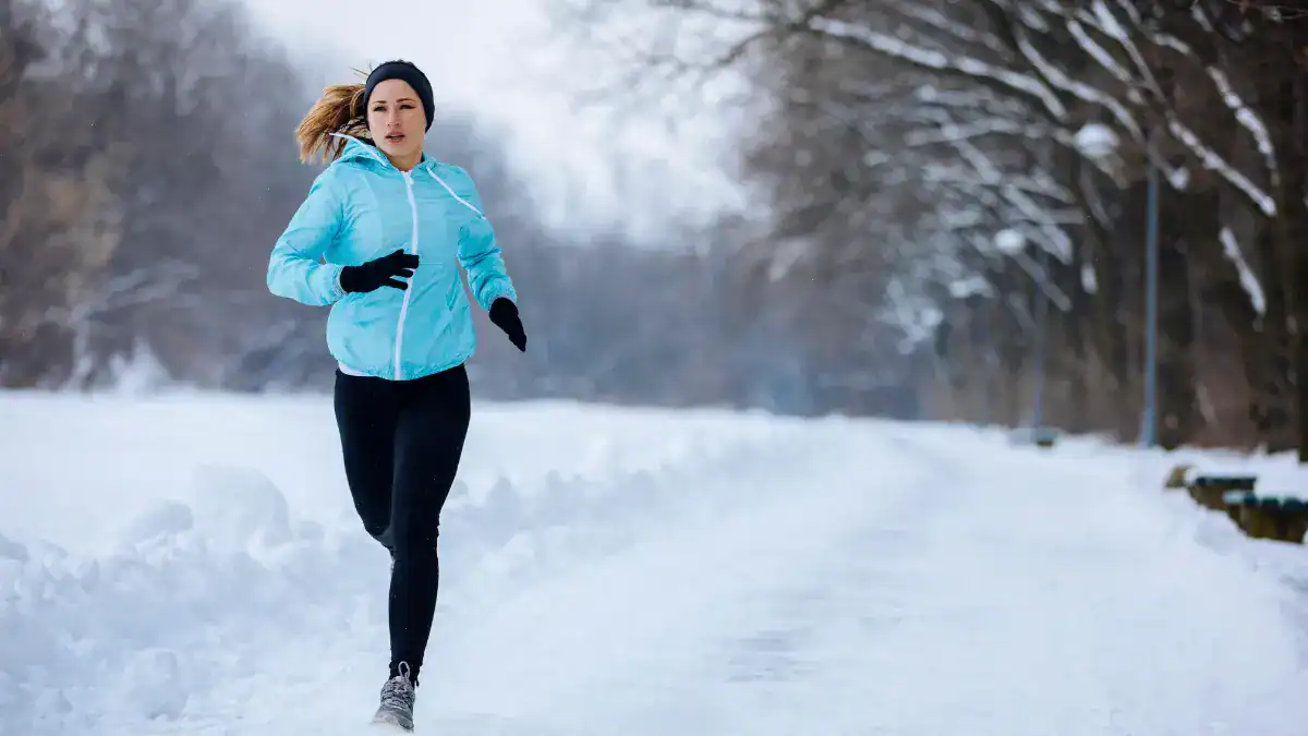 A person wearing a light blue jacket, black leggings, gloves, and a headband runs on a snow-covered path through a winter landscape.
