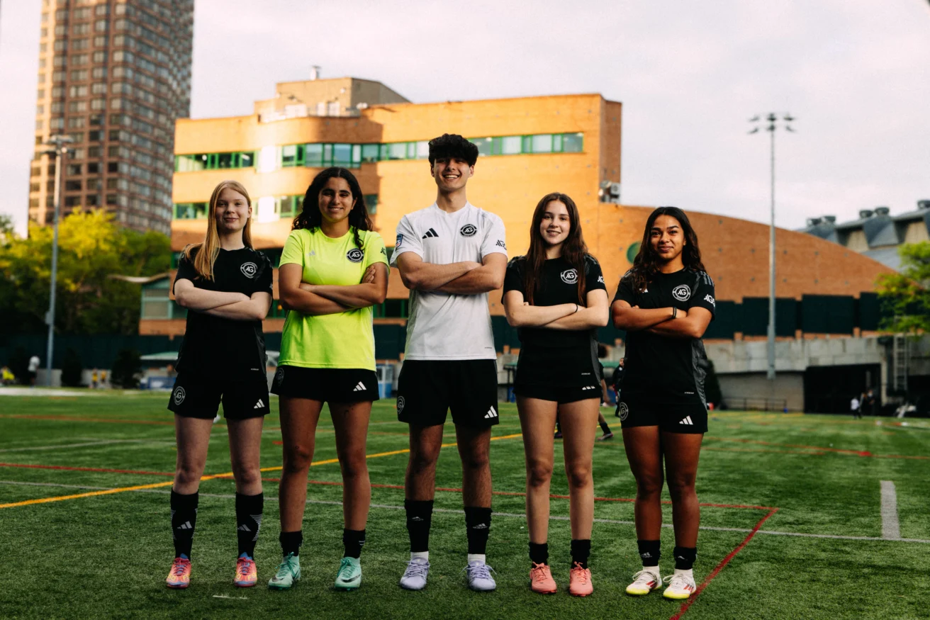 Five AGSC youth soccer players stand in a row with arms crossed on an outdoor field, wearing matching black uniforms, with a city building in the background.