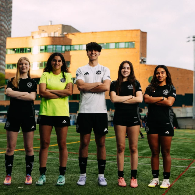 Five AGSC youth soccer players stand in a row with arms crossed on an outdoor field, wearing matching black uniforms, with a city building in the background.