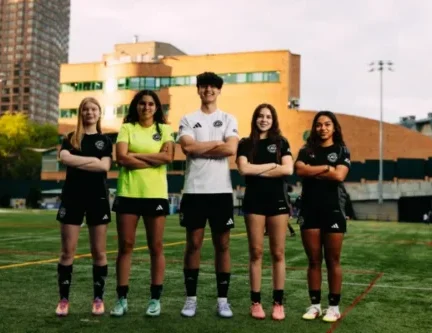 Five AGSC youth soccer players stand in a row with arms crossed on an outdoor field, wearing matching black uniforms, with a city building in the background.