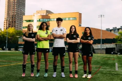 Five AGSC youth soccer players stand in a row with arms crossed on an outdoor field, wearing matching black uniforms, with a city building in the background.