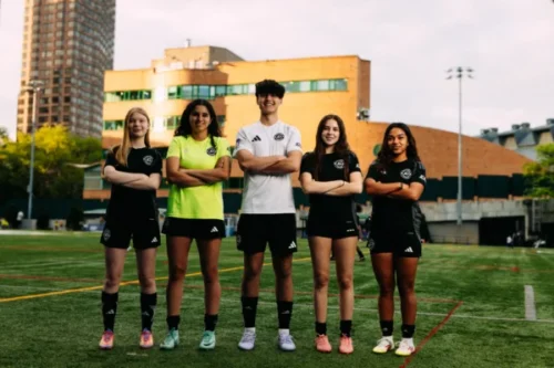 Five AGSC youth soccer players stand in a row with arms crossed on an outdoor field, wearing matching black uniforms, with a city building in the background.