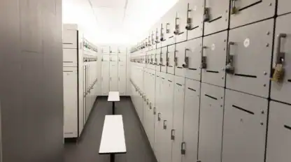 A row of white lockers with combination and padlocks lines both sides of a narrow room, with two white benches in the center under ceiling lights.