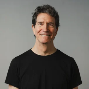 A middle-aged man with graying hair smiles at the camera. He is wearing a plain black t-shirt and standing against a neutral gray background.