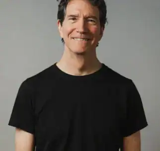 A middle-aged man with graying hair smiles at the camera. He is wearing a plain black t-shirt and standing against a neutral gray background.