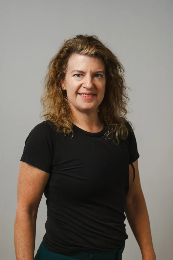 A woman with wavy, shoulder-length brown hair wearing a black t-shirt stands against a plain light gray background, facing the camera and smiling.