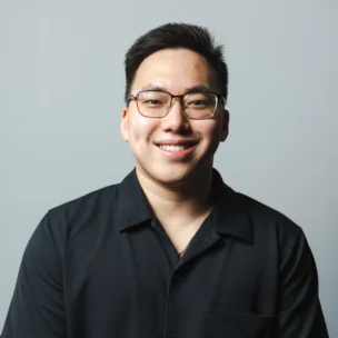 A young man with short dark hair, glasses, and a black button-up shirt smiles at the camera against a plain light grey background.