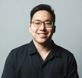 A young man with short dark hair, glasses, and a black button-up shirt smiles at the camera against a plain light grey background.