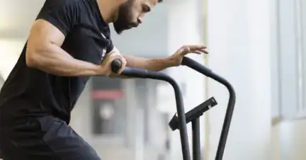 A man wearing a black shirt and shorts uses an exercise bike indoors, holding the handlebars and looking at the display.