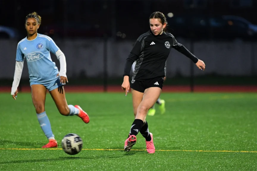 Two female soccer players compete on a field, with one in a black uniform kicking the ball and the other in a light blue uniform running nearby.