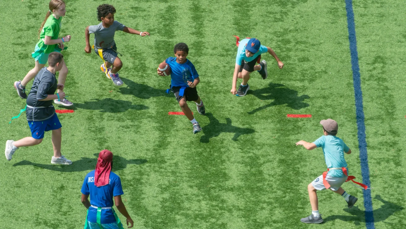 Six children play flag football on a green field, with one child running with the ball while others attempt to grab his flag.