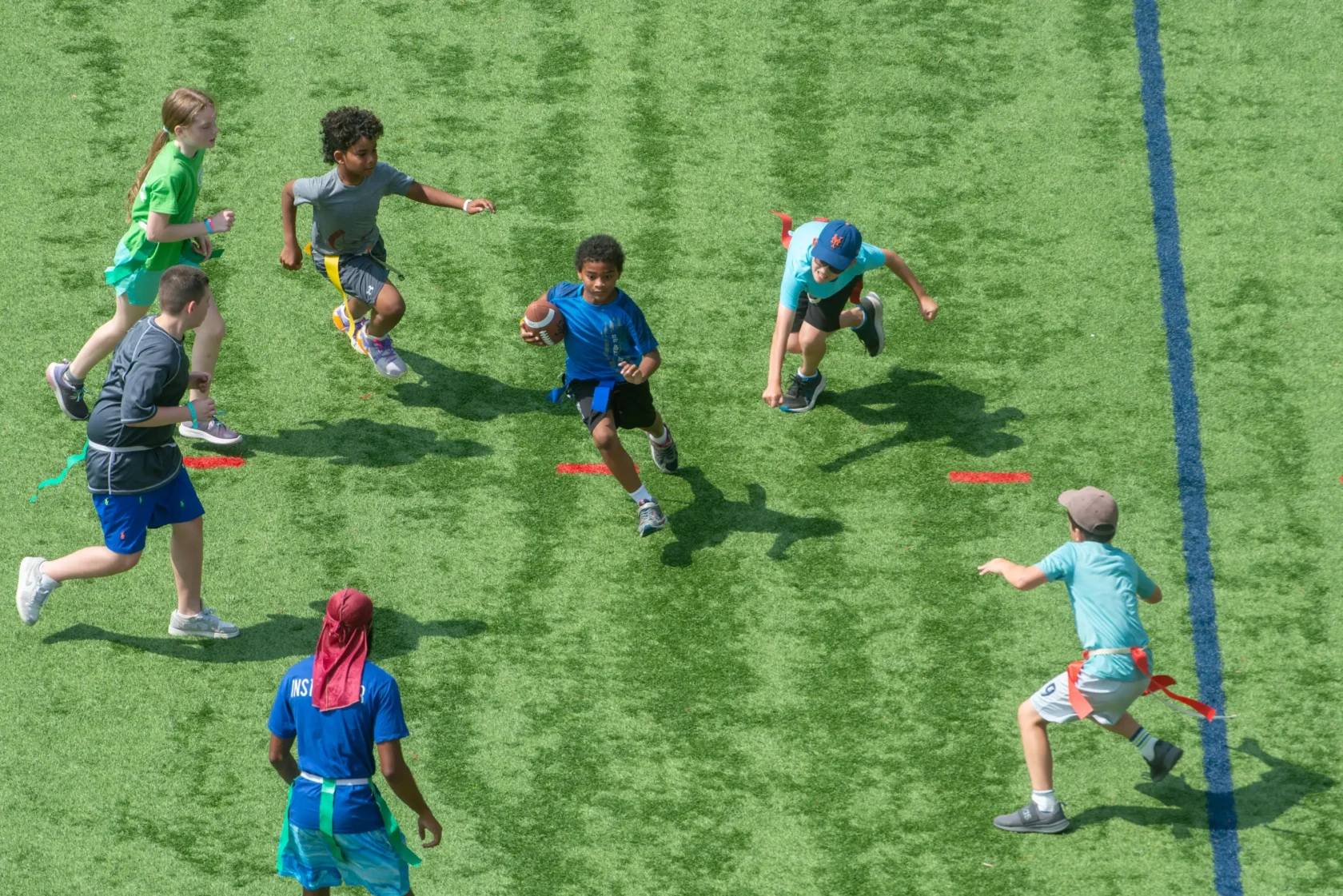 Six children play flag football on a green field, with one child running with the ball while others attempt to grab his flag.