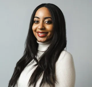 A woman with long dark hair wearing a white ribbed turtleneck sweater stands against a plain light gray background, smiling at the camera.