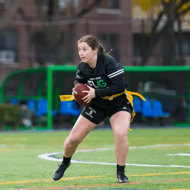 A young woman holding a football prepares to throw during a flag football game on an outdoor field.