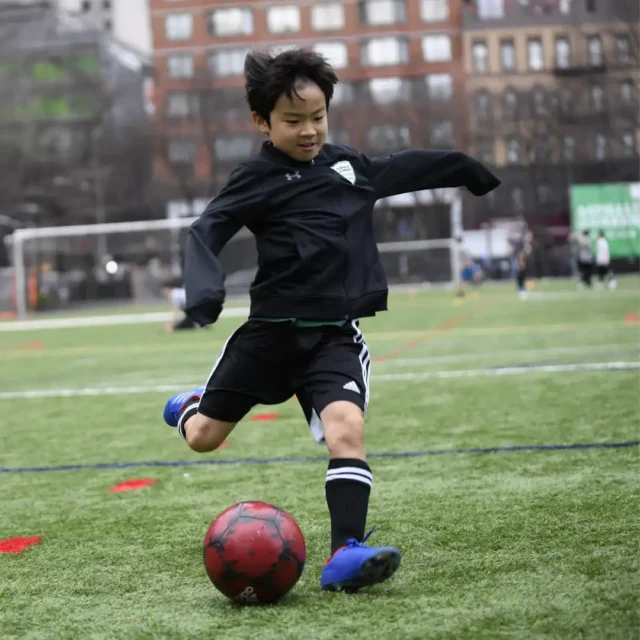 A child in sportswear is about to kick a red soccer ball on an outdoor soccer field, with buildings and other players in the background.