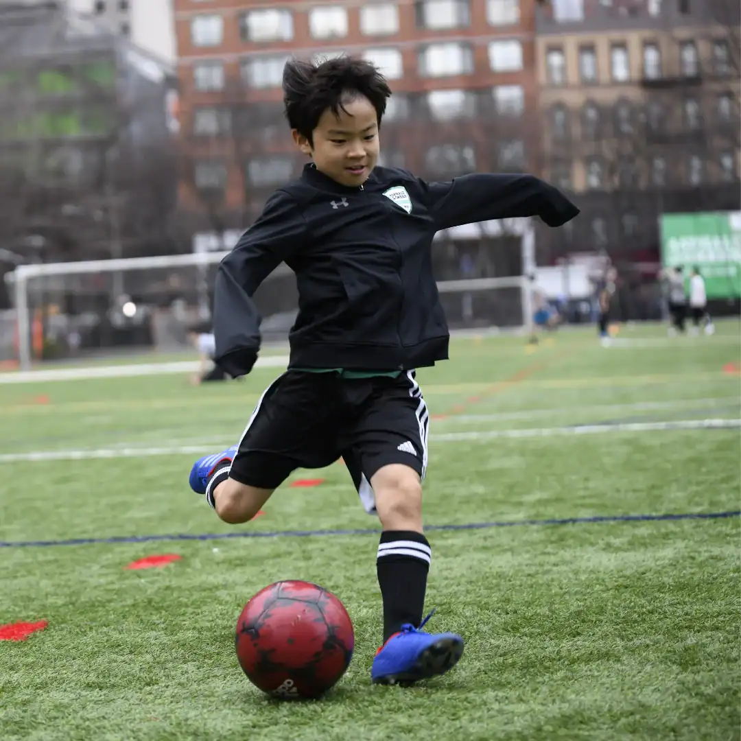 A child in sportswear is about to kick a red soccer ball on an outdoor soccer field, with buildings and other players in the background.