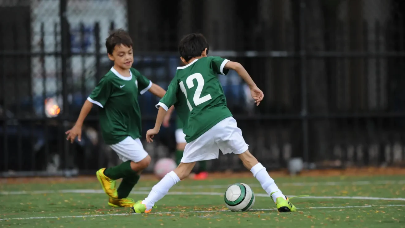Two young boys in green soccer uniforms play on a field; one dribbles the ball while the other runs nearby.