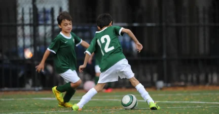 Two young boys in green soccer uniforms play on a field; one dribbles the ball while the other runs nearby.