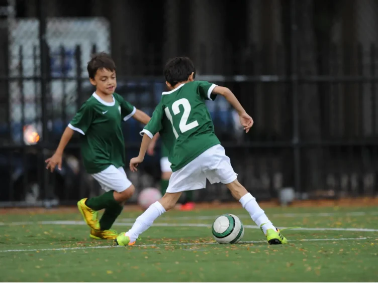 Two young boys in green soccer uniforms play on a field; one dribbles the ball while the other runs nearby.