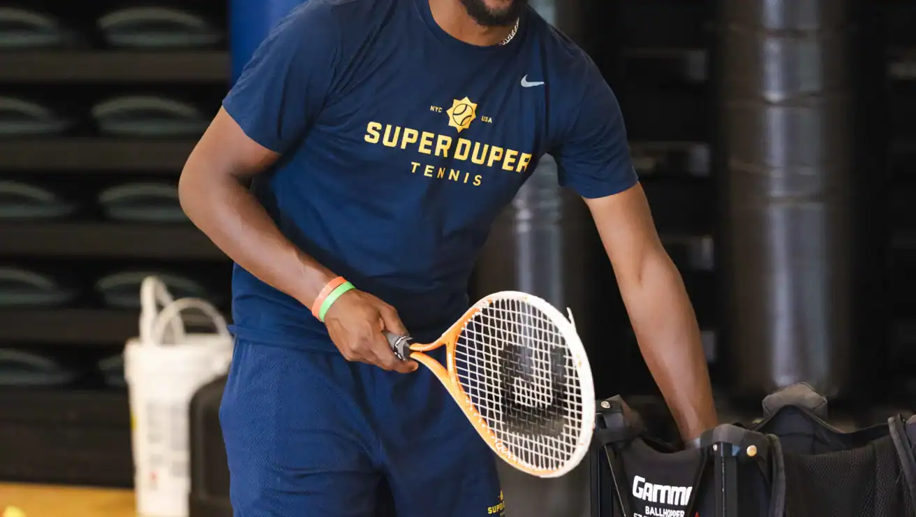 A tennis coach wearing a "SuperDuper Tennis" shirt smiles while holding a racket and standing next to a basket of tennis balls in an indoor gym.