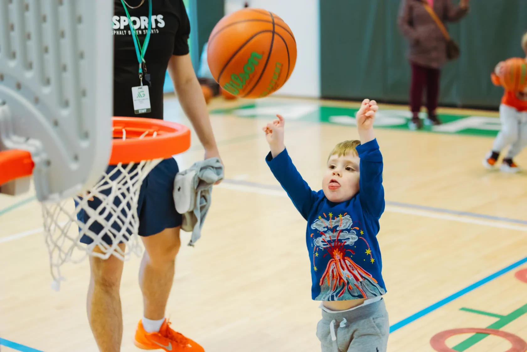 A young child in a blue shirt attempts a basketball shot toward a hoop in a gym, with an adult and other children in the background.