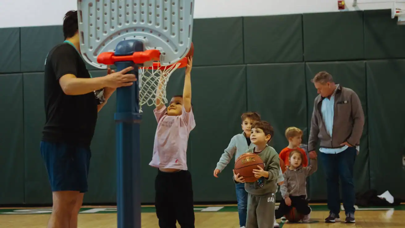 A group of young children play basketball indoors; one child hangs from the hoop, while others, along with two adults, stand nearby on the gym floor.