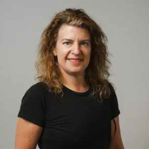 A woman with wavy brown hair wearing a black t-shirt stands in front of a plain light grey background, looking at the camera and smiling.