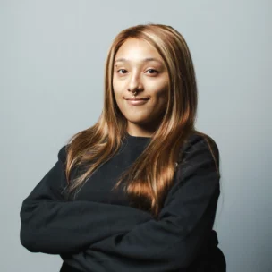 Person with long, straight light brown hair and a nose ring, wearing a black sweater, stands with arms crossed against a plain light gray background.
