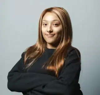 Person with long, straight light brown hair and a nose ring, wearing a black sweater, stands with arms crossed against a plain light gray background.