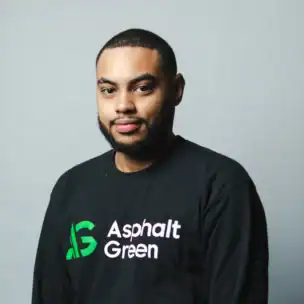 A man with short hair and a beard wearing a black "Asphalt Green" shirt stands against a plain light gray background.