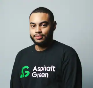 A man with short hair and a beard wearing a black "Asphalt Green" shirt stands against a plain light gray background.