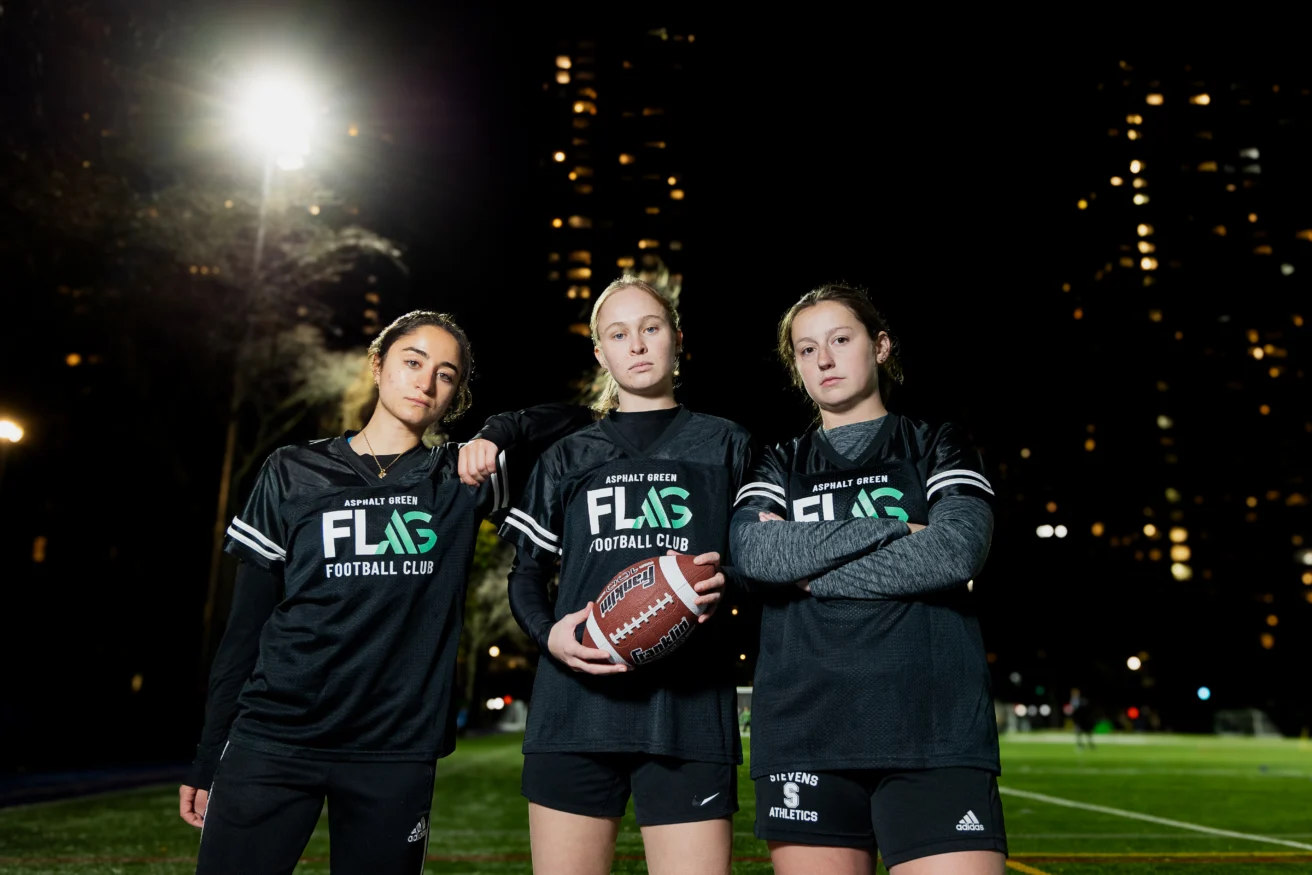 Three women in black "Flag Football Club" jerseys pose on a lit outdoor football field at night, with city buildings visible in the background. One holds a football.