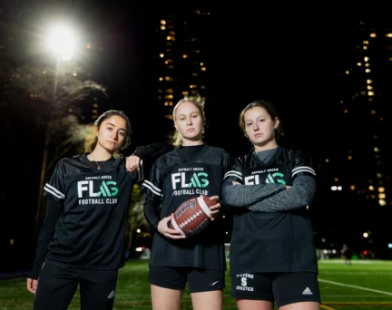 Three women in black "Flag Football Club" jerseys pose on a lit outdoor football field at night, with city buildings visible in the background. One holds a football.