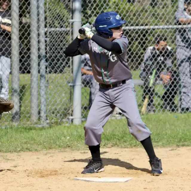 A youth baseball player in a gray uniform stands in the batter’s box, holding a bat and preparing to hit, with a chain-link fence and teammates in the background.