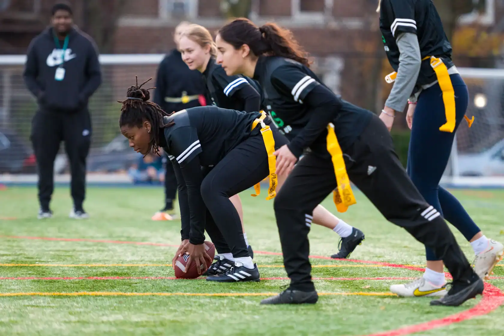 A group of women prepare for a play during a youth flag football game on an outdoor field, with a coach or referee observing in the background.