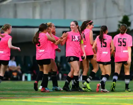 A group of AGSC girls players jerseys celebrate together on a soccer field during a game.