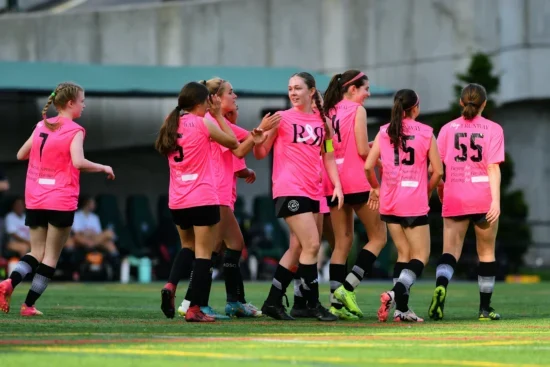A group of AGSC girls players jerseys celebrate together on a soccer field during a game.