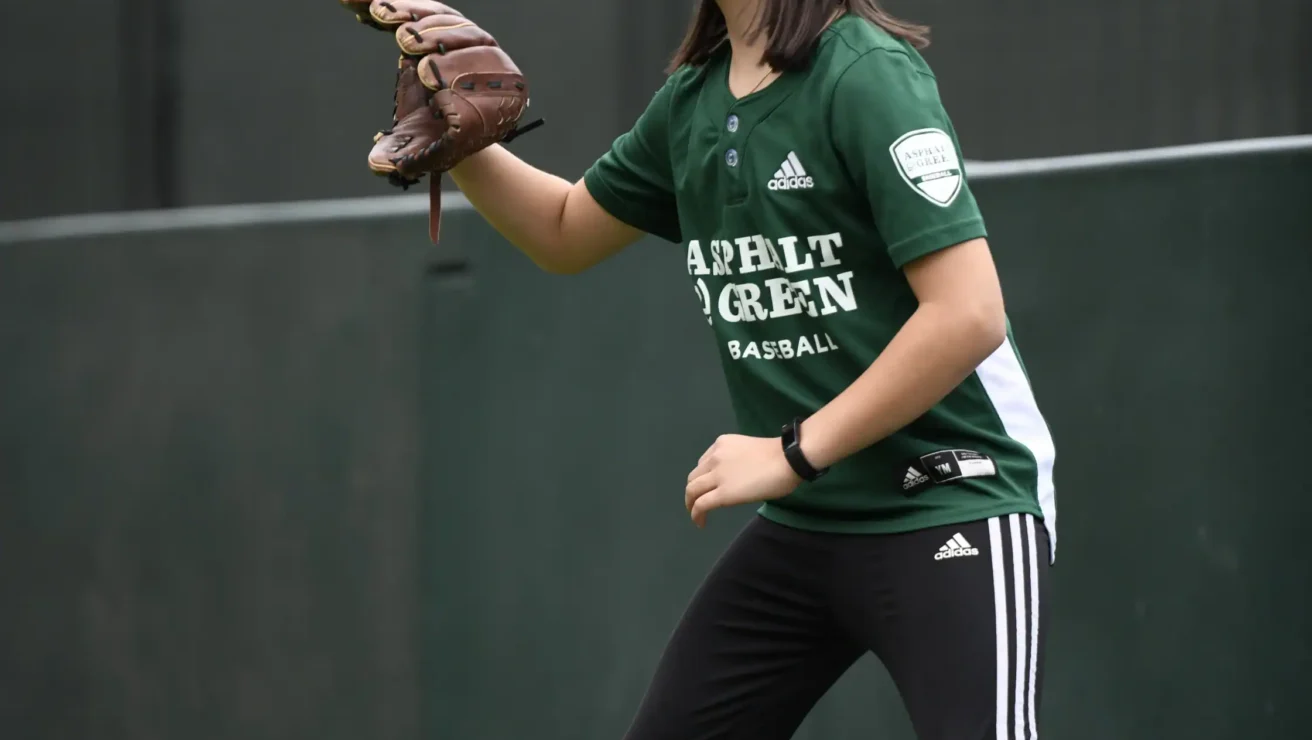 A young athlete in a green jersey and black hat stands ready with a glove, preparing to catch a ball on a youth baseball field.
