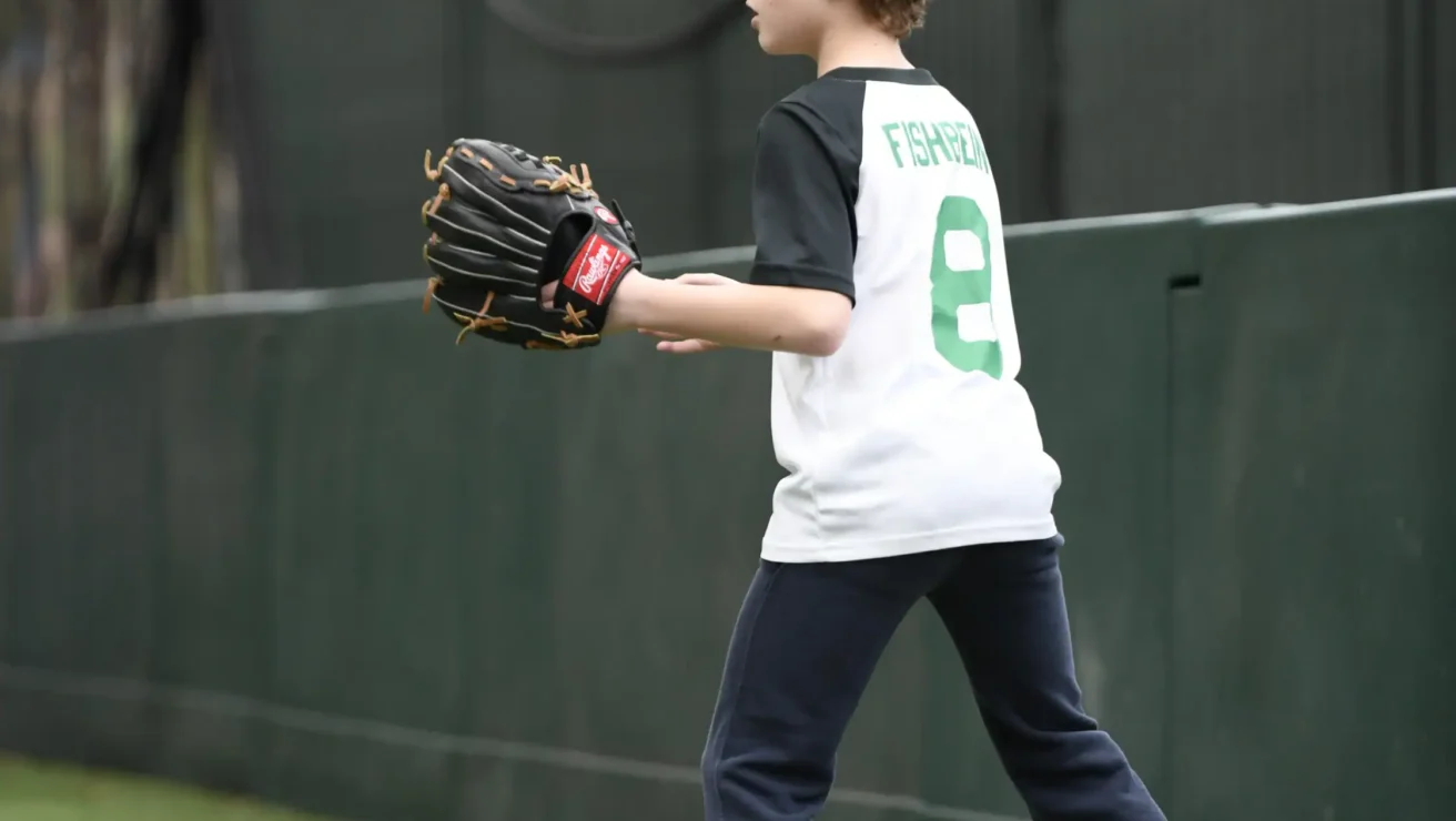 A young boy wearing a baseball glove stands on artificial grass near a fence, ready to catch a ball during youth baseball practice.